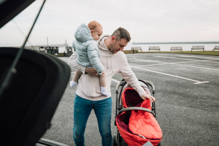 Male foster carer holding a toddler and lifting a folder pushchair into the boot of his car