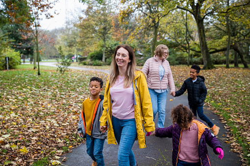 Front-view shot of a multi-gen fostering family walking along a footpath in a public park, autumn leaves cover the ground, they are wearing winter clothing while holding hands and smiling.
