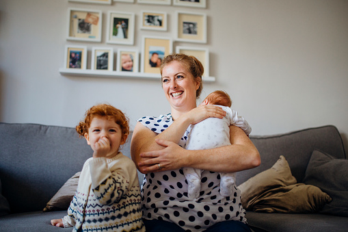 Foster carer with two children in the living room of their home. The mother is holding a baby and the other little girl is laughing and looking at the camera while sucking her thumb.